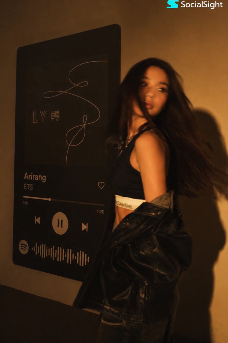 一位发型飘逸的深色长发时尚年轻女子在巨型壁挂Spotify音乐播放器显示屏前摆姿势。温暖的琥珀色电影级灯光布满房间，在墙上投下戏剧性阴影。她身穿黑色卡尔文·克莱因露脐上衣，廓形黑色皮夹克滑落肩...