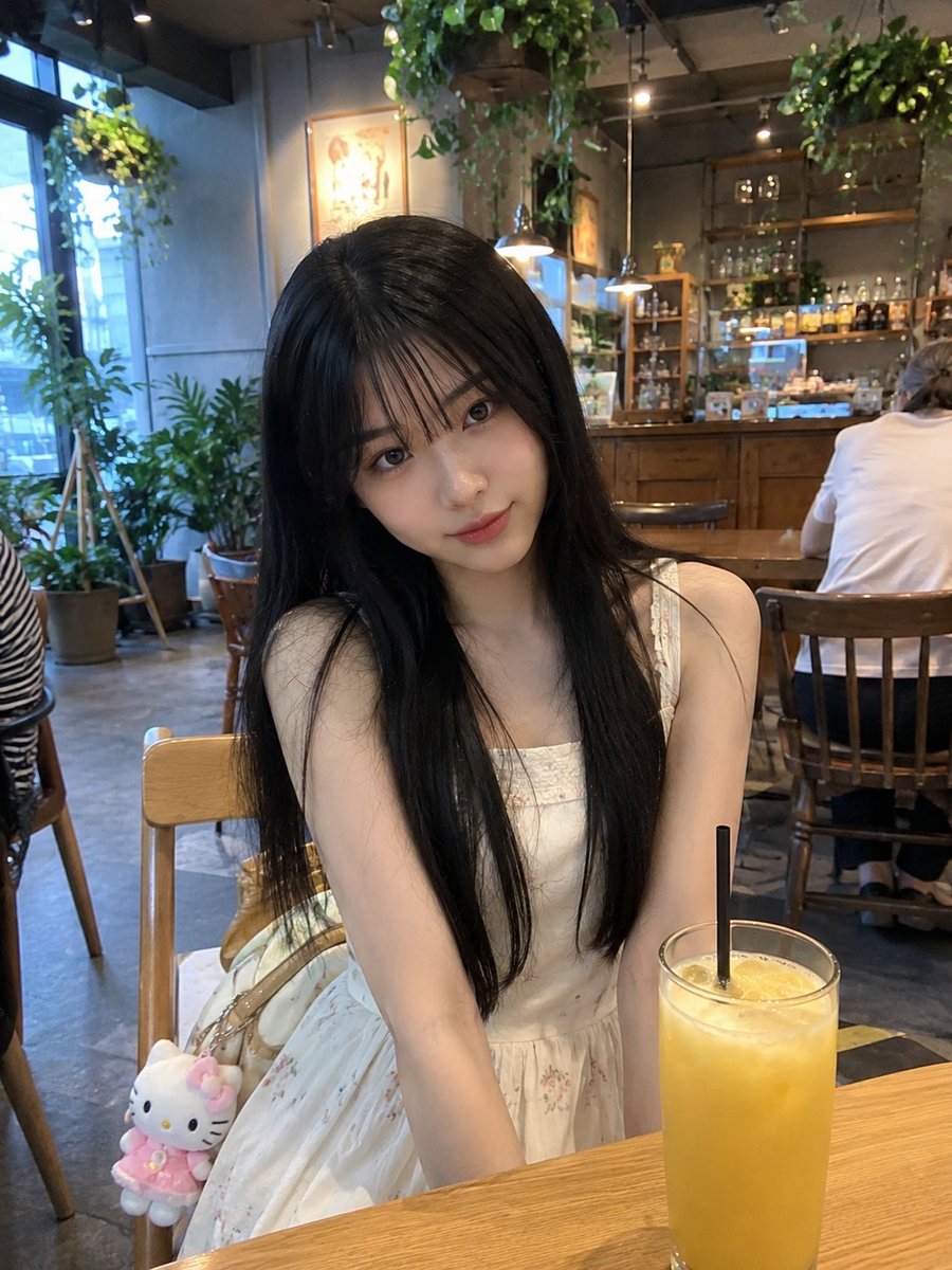 年轻的日本女孩，留着柔顺的长黑发、轻柔蓬松的刘海、白皙透亮的肌肤和一双大而传神的眼睛，随意地坐在一家温馨现代的咖啡馆里。身穿一件精致的奶油白无袖连衣裙，裙身点缀着细腻的花卉图案，双手自然地放在...