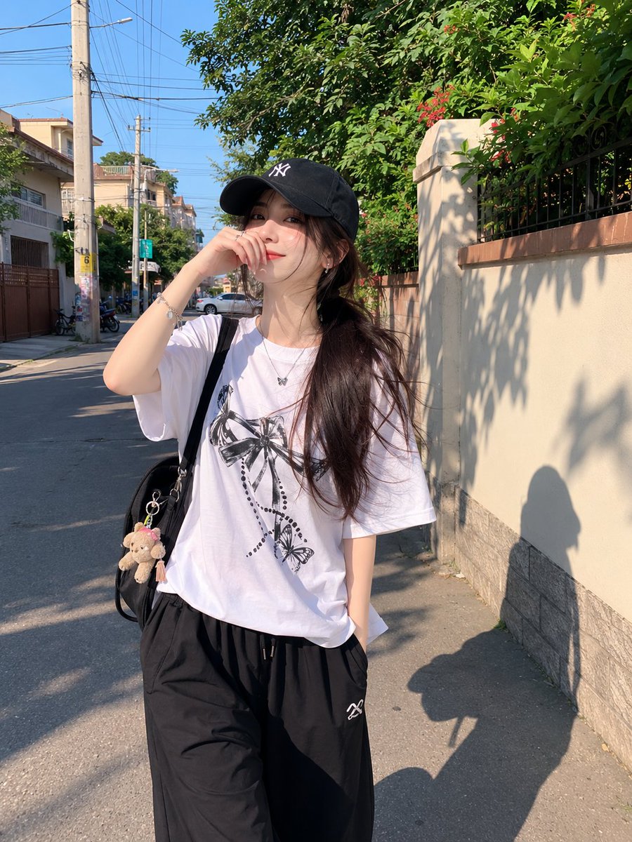 年轻漂亮的日本女孩，肤色白皙，妆容自然柔和，长长的深色头发在黑色棒球帽下扎成低马尾，身穿宽松的白色印花T恤和黑色宽松全长运动裤。在一个晴朗的下午，她随意地走过一条安静的住宅小巷，一只手自然地举...