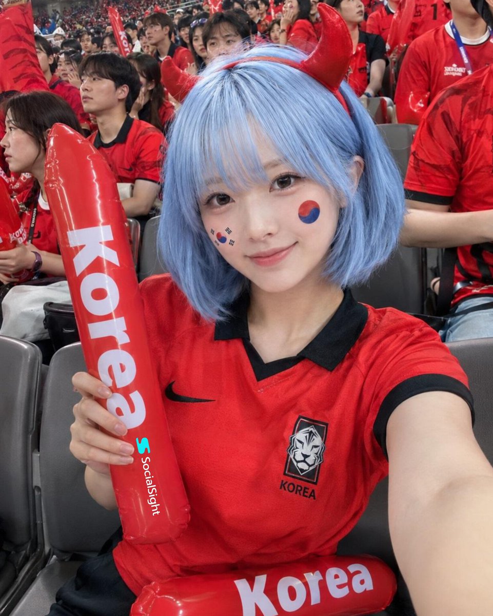 在一座座无虚席的体育场内，国际足球比赛期间，一位美丽的足球迷坐在看台上拍摄的极致写实自拍，齐耳短款粉蓝色波波头，闪亮的红色恶魔角发箍，柔和自然妆容，明亮有神的眼睛，对着镜头露出淡淡的微笑。身穿...