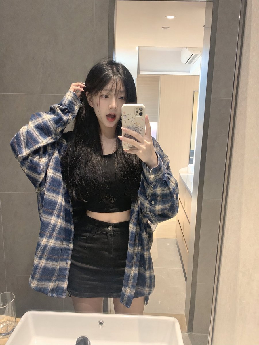 留着长长的黑色直发和柔软稀疏刘海的年轻日本女孩，内穿黑色短款露脐上衣，外穿 oversized 蓝色格纹法兰绒衬衫，搭配黑色迷你短裙。站在浴室镜子前，拿着配有可爱卡通保护壳的智能手机，正在拍一...