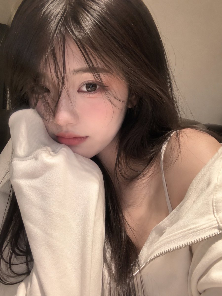 一张照片般逼真的特写自拍，一位可爱的年轻女性，脸部占据画面的大部分，头部微微倾斜，脸颊舒适地靠在一件宽松的奶油色连帽衫袖子里。柔顺的长发配有柔和的八字刘海遮住一只眼睛，大而有神的眼睛带着上挑的...
