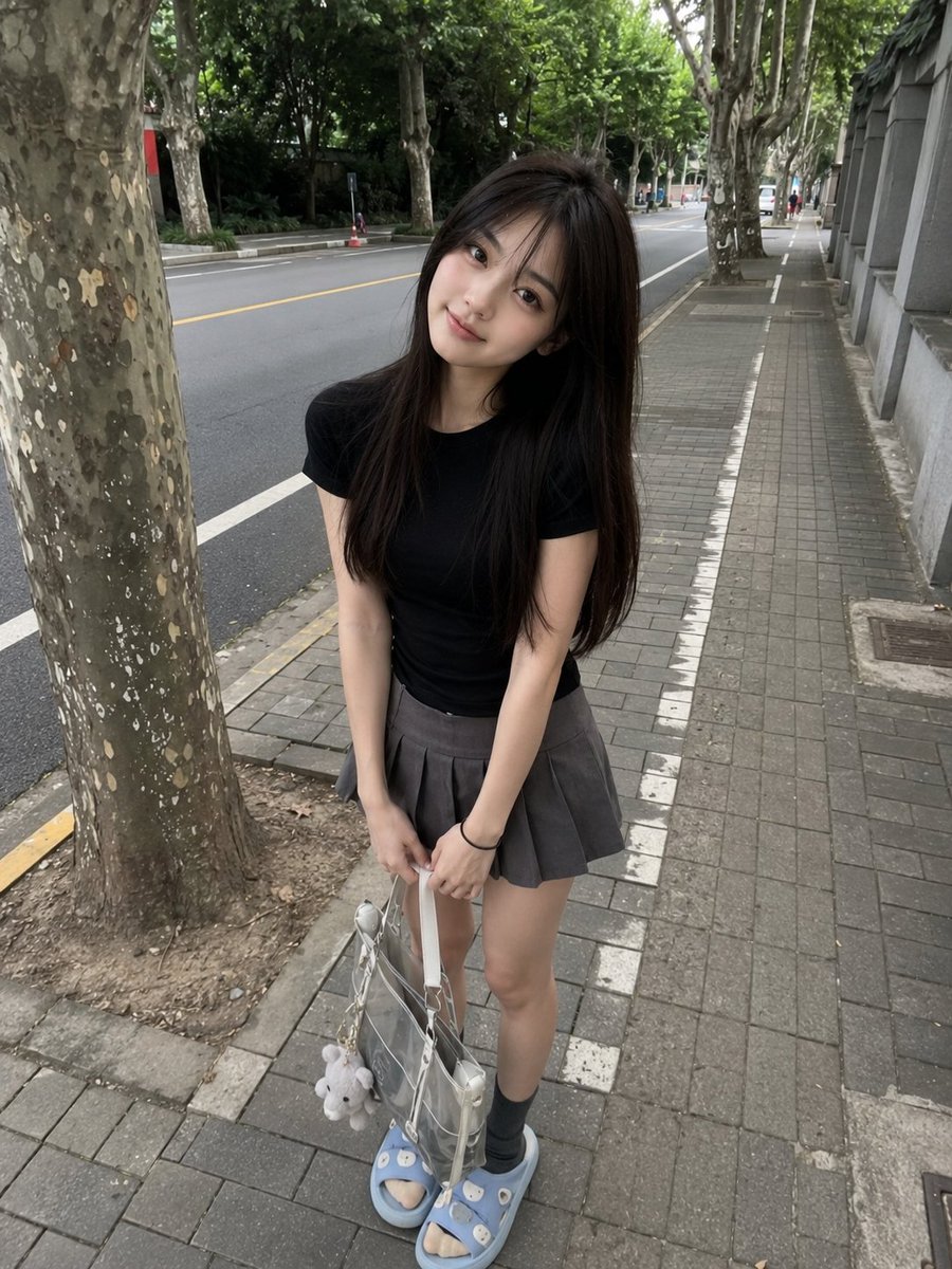 年轻的日本女孩，长直深棕色头发配柔和刘海，身着修身黑色T恤、灰色百褶迷你裙、灰色袜子与浅蓝色拖鞋，随意站在绿树成行的安静道路旁的人行道上。微微前倾，露出可爱自然的微笑，手里拿着一个透明托特包，...