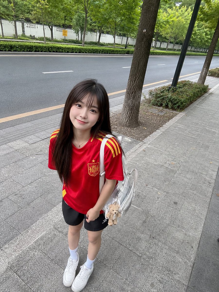 年轻的日本女孩，留着长直的深棕色头发和柔软的刘海，身穿红色西班牙国家足球队球衣、黑色运动短裤、白色中筒袜和白色运动鞋，悠闲地站在绿树成荫的安静道路旁的人行道上。她微微前倾，露出可爱自然的微笑，...