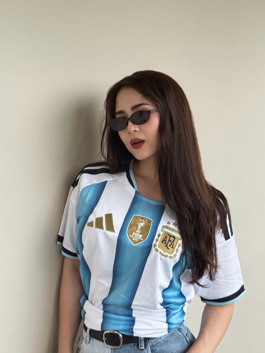 超现实风格的时尚人像摄影：一位时髦的年轻女性，身穿不同国家队的足球球衣（阿根廷、葡萄牙、巴西），在室内一面干净极简的米白色墙壁前自信地摆姿势。佩戴时髦的窄框黑色太阳镜，肌肤白皙无瑕，自然妆容配...