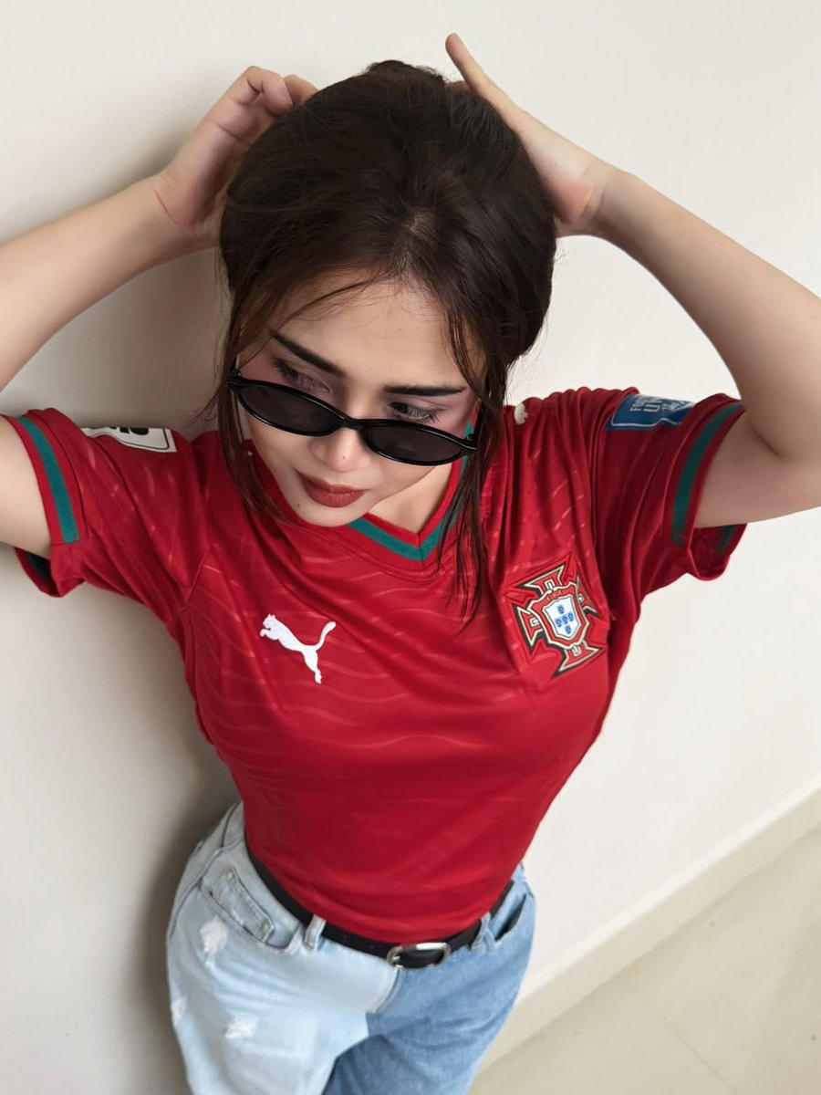 超现实风格的时尚人像摄影：一位时髦的年轻女性，身穿不同国家队的足球球衣（阿根廷、葡萄牙、巴西），在室内一面干净极简的米白色墙壁前自信地摆姿势。佩戴时髦的窄框黑色太阳镜，肌肤白皙无瑕，自然妆容配...