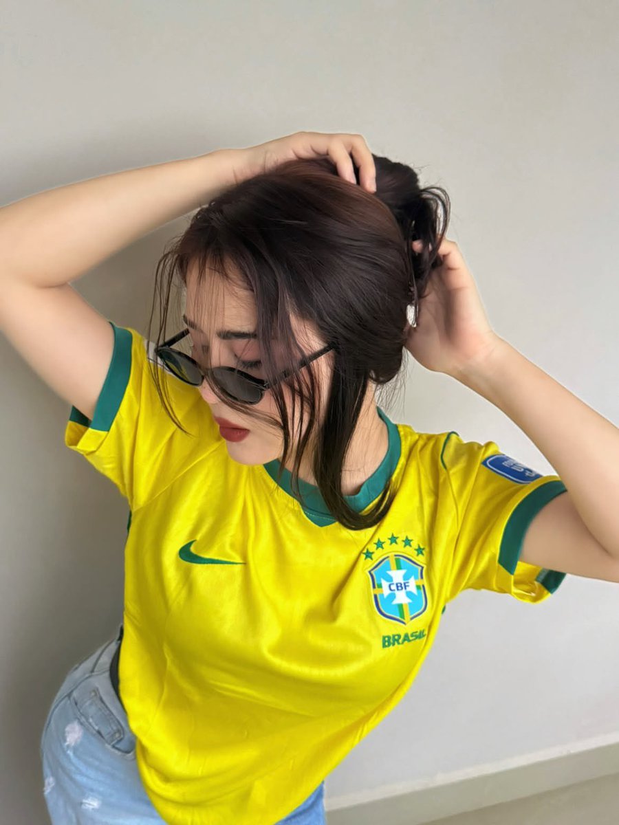 超现实风格的时尚人像摄影：一位时髦的年轻女性，身穿不同国家队的足球球衣（阿根廷、葡萄牙、巴西），在室内一面干净极简的米白色墙壁前自信地摆姿势。佩戴时髦的窄框黑色太阳镜，肌肤白皙无瑕，自然妆容配...