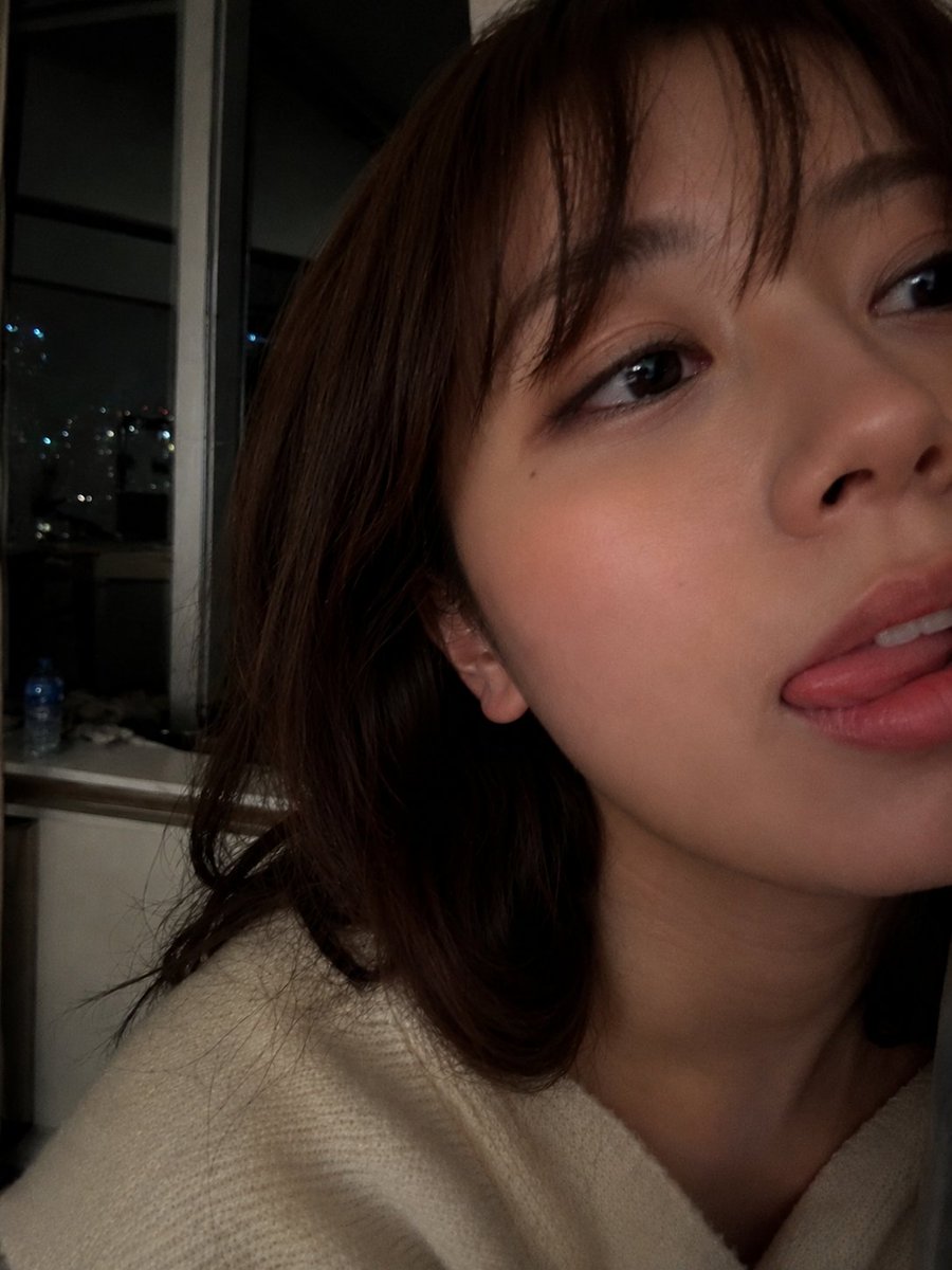 室内夜景中，使用智能手机摄像头拍摄的年轻女性超写实特写自拍肖像。肌肤柔和自然，肤质光滑细腻，双颊微微泛红，嘴唇粉嫩水润，表情俏皮，舌头轻触上唇。一双深色的大眼睛略带灵动，微微侧向镜头方向。中长...