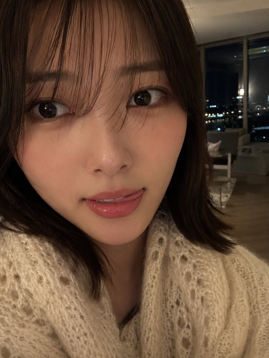 室内夜景中，使用智能手机摄像头拍摄的年轻女性超写实特写自拍肖像。肌肤柔和自然，肤质光滑细腻，双颊微微泛红，嘴唇粉嫩水润，表情俏皮，舌头轻触上唇。一双深色的大眼睛略带灵动，微微侧向镜头方向。中长...