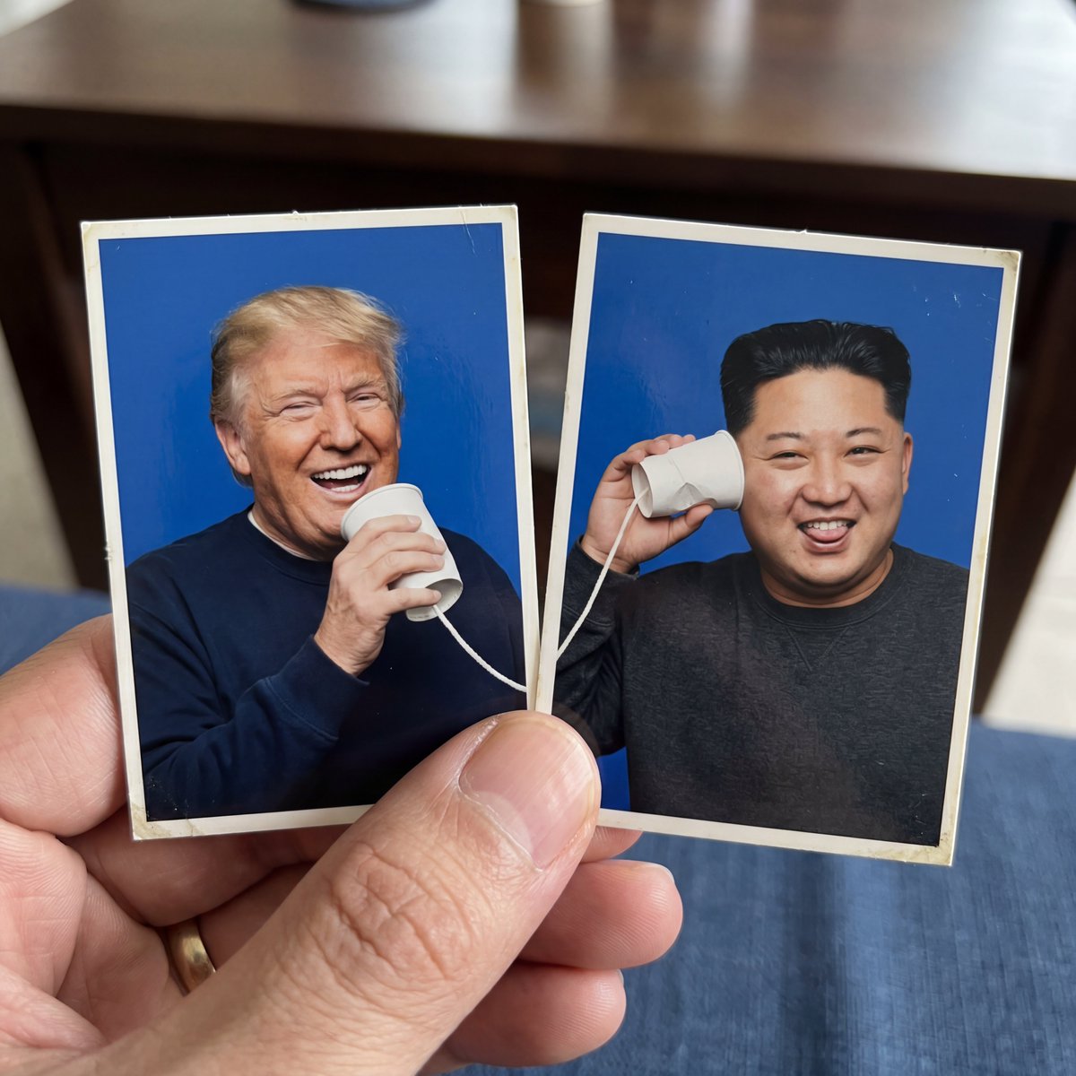 创作一个创意1:1正方形构图，包含两张并排摆放的护照式照片打印件，被一只手的拇指和食指捏住。使用上传的参考图像作为两个拍摄对象，准确保留每个对象的面部特征、发型、身份和整体外观。两张护照照片都...