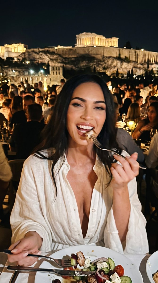 一张微妙模糊、低画质的特写业余 iPhone 照片，内容是一位惊艳的年轻 Megan Fox（梅根·福克斯），在雅典帕特农神庙附近一家高档拥挤的餐厅里，身穿一袭全白色女神式未扣纽扣的连衣裙，一...