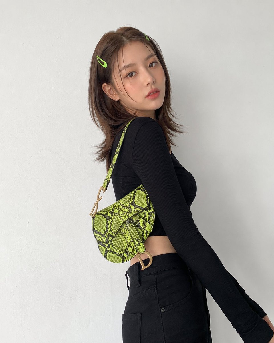 创建一张图片，画面中有一位年轻的韩国女性，留着中长棕色头发，两侧别着青柠绿色的发夹，背靠一面纯白色的墙壁摆姿势，身穿黑色长袖短款上衣、黑色高腰长裤，肩上斜挎一只颜色鲜艳的青柠绿色蛇纹马鞍包。