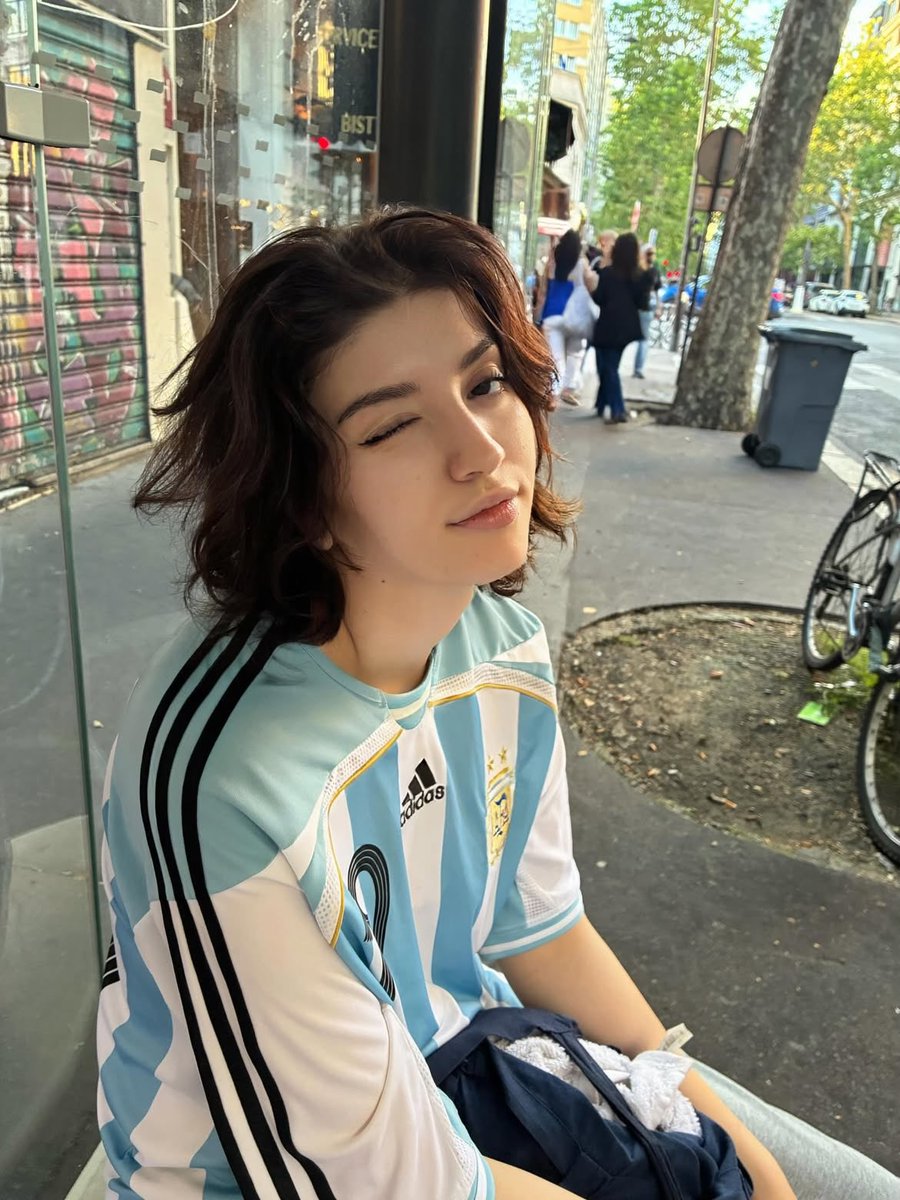 超写实的随性人像摄影：一位留着短波浪深色头发的年轻人，身着阿根廷国家足球队宽大球衣（19号），在黄金时段坐在城市公交车内和公交车站上。柔和温暖的阳光透过大窗户洒进来，在脸上形成戏剧性的明暗光影...