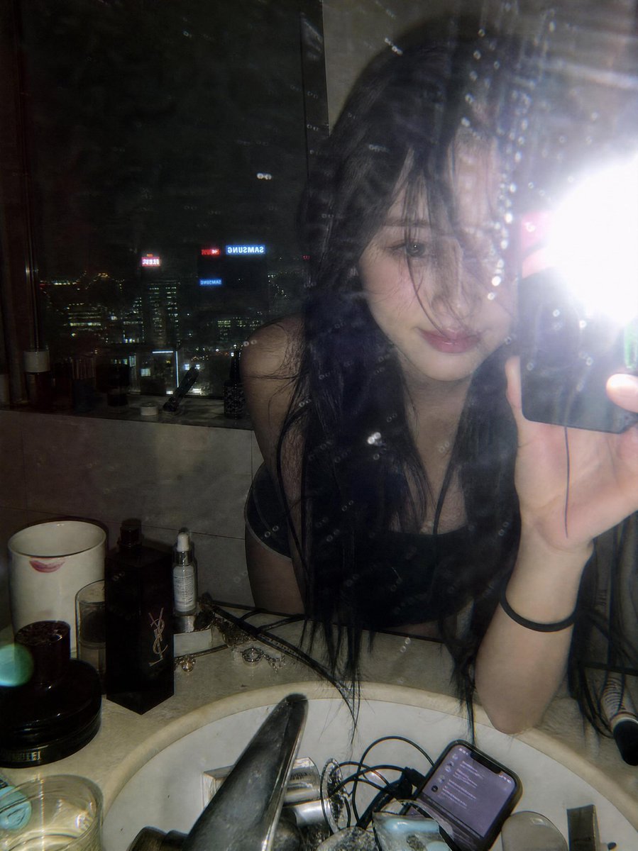 一张氛围感十足的深夜镜面自拍，拍摄于一间俯瞰璀璨城市天际线的豪华高层公寓浴室中。一位年轻女性留着略带凌乱黑色长发，身体向前倚靠在白色大理石洗手台上方，蓬松的发丝若隐若现地遮挡着一只眼睛。照片透...