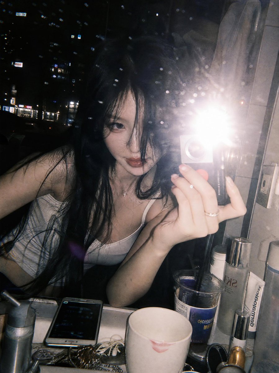 一张氛围感十足的深夜镜面自拍，拍摄于一间俯瞰璀璨城市天际线的豪华高层公寓浴室中。一位年轻女性留着略带凌乱黑色长发，身体向前倚靠在白色大理石洗手台上方，蓬松的发丝若隐若现地遮挡着一只眼睛。照片透...