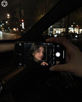照片于夜间在车内拍摄；画面中仅露出一只女性的手和一部iPhone，年轻女性的面部仅出现在手机屏幕上。相机位于副驾驶侧，构图同时纳入挡风玻璃与手机，手机被单手直接握在人物前方。手中握着一部横向持...