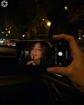 照片拍摄于夜间车内；画面中仅可见女性的一只手和一部iPhone，而年轻女性的面部仅出现在手机屏幕上。相机位于副驾驶一侧，取景框对准挡风玻璃和手机，手机由单手举起，置于人物正前方。手中握着一台横...