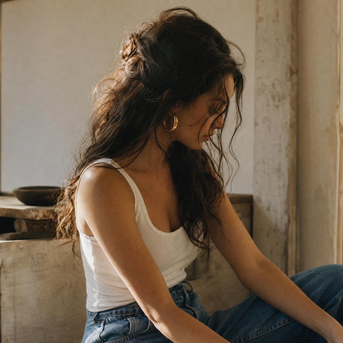 一张自然随性的中景人像照：一位女性拥有蓬松凌乱、波浪卷曲的深棕色长发，部分以半扎发样式向后别起，鬓角处有几缕碎发自然垂落，修饰着脸部轮廓。她若有所思地向下凝视，画面以柔和、温暖的侧脸角度捕捉。...