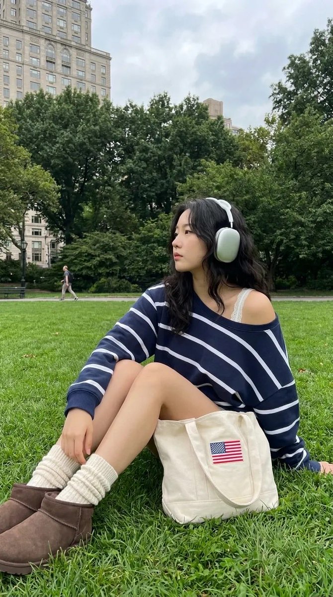 中景全身镜头：一位时髦的24岁韩国女性在像中央公园这样的知名城市公园的翠绿草坪上放松休憩。她弯曲膝盖坐着，身体微微后倾，姿态放松，同时转头看向镜头外，露出宁静沉思的侧脸。她佩戴着一副现代风格的...
