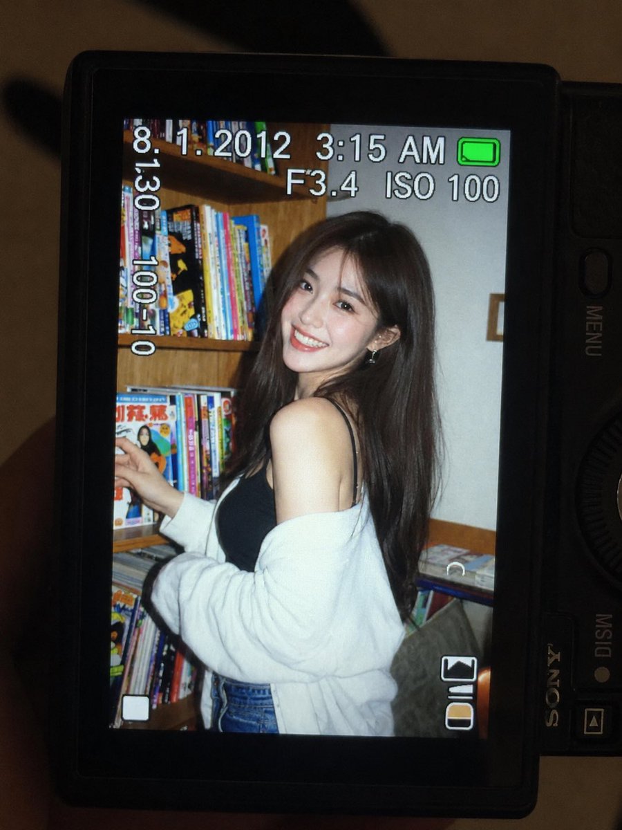 一张超精细的特写镜头：一台微型紧凑型数码相机的 LCD 屏幕在原本黑暗的室内房间中明亮发光。屏幕上是一张 2010 年代初期的随性傻瓜相机快照，画面中一位年轻东亚女性留着一头深色长波浪卷发，站...