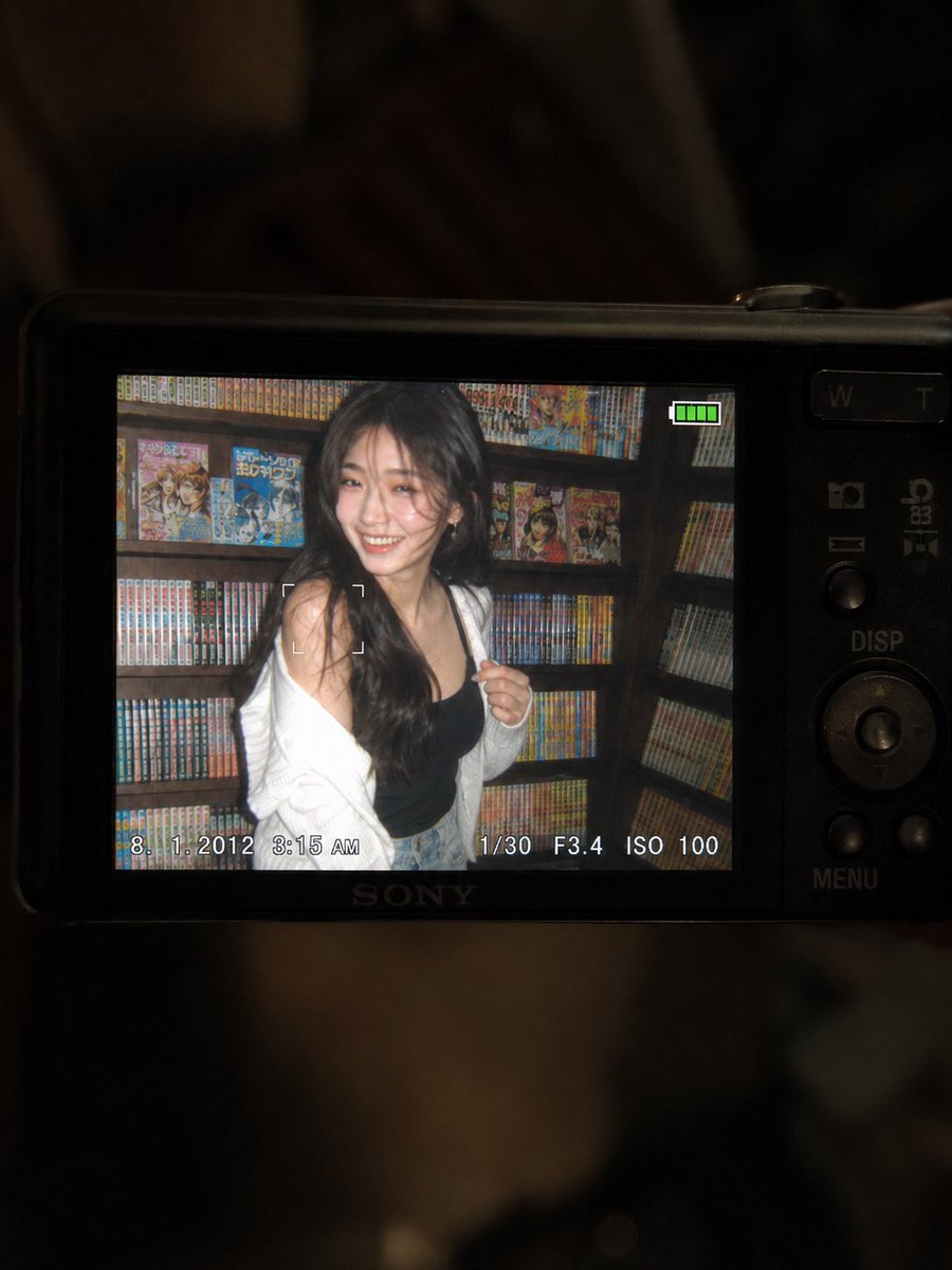 一张超精细的特写镜头：一台微型紧凑型数码相机的 LCD 屏幕在原本黑暗的室内房间中明亮发光。屏幕上是一张 2010 年代初期的随性傻瓜相机快照，画面中一位年轻东亚女性留着一头深色长波浪卷发，站...