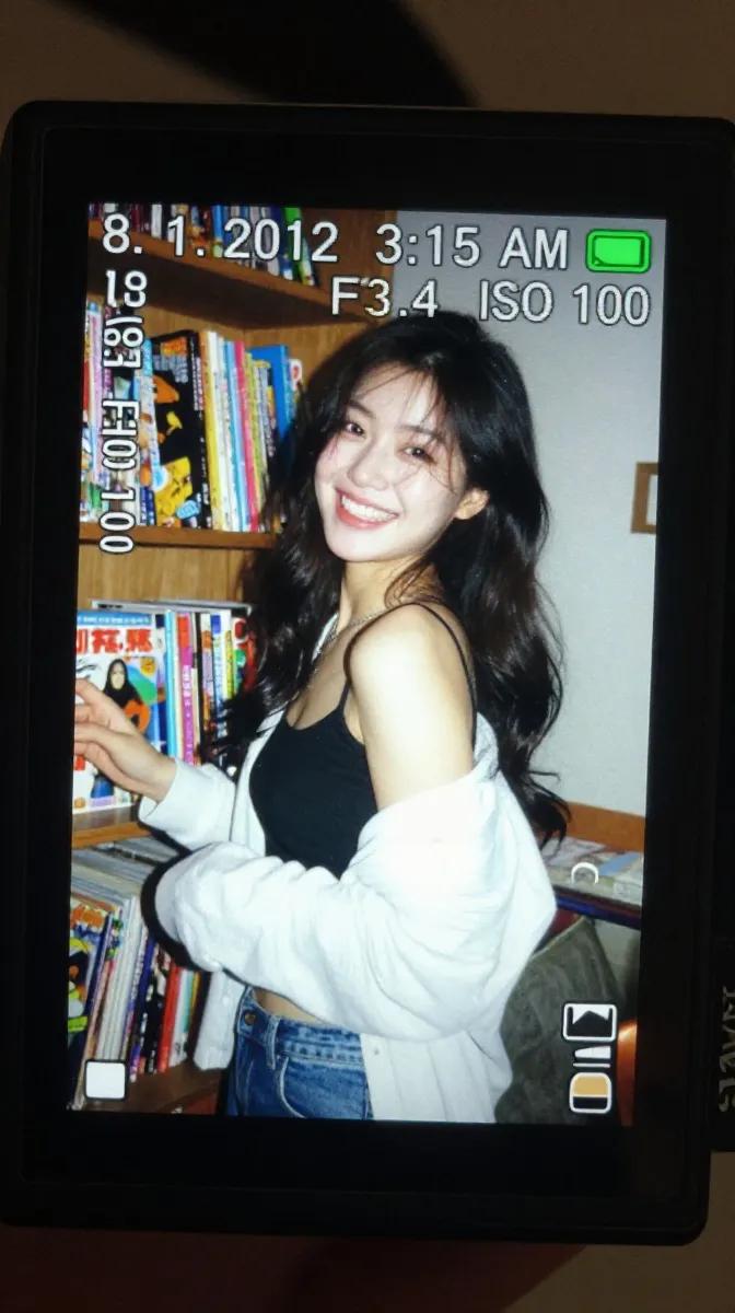 一个真实感的特写镜头，拍摄的是在昏暗室内环境中明亮发光的一台小型数码相机屏幕。LCD 屏幕上显示着一张抓拍风格的早期 2010 年代风格照片，照片中一位年轻东亚女性留着长长的深色波浪卷发，站在...