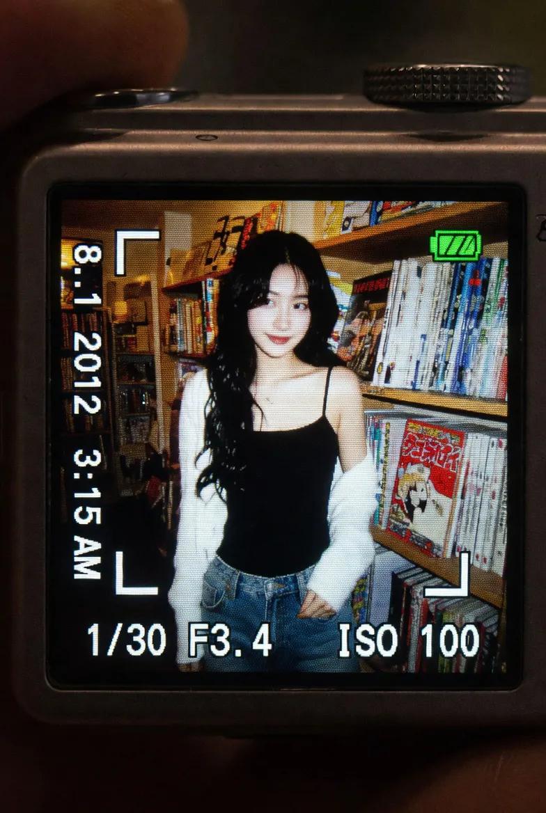 一个真实感的特写镜头，拍摄的是在昏暗室内环境中明亮发光的一台小型数码相机屏幕。LCD 屏幕上显示着一张抓拍风格的早期 2010 年代风格照片，照片中一位年轻东亚女性留着长长的深色波浪卷发，站在...