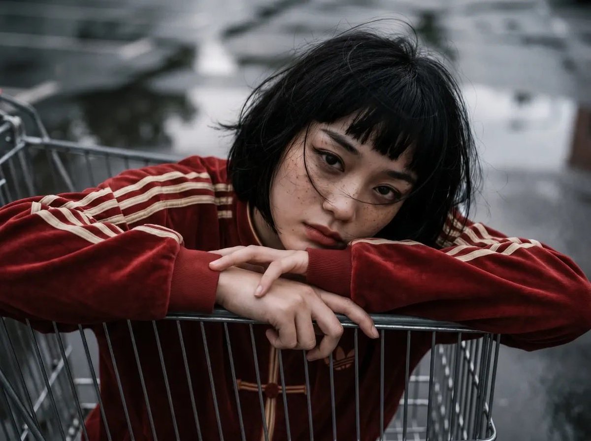 女性角色位于购物推车内部，以推车内部为视角向上望向她的脸部，戏剧性的透视畸变，一条腿靠近镜头，姿势"无所谓"且放松：，超大红色（偏红）的阿迪达斯旗袍风格天鹅绒运动套装，配米色条纹与盘扣，搭配同...