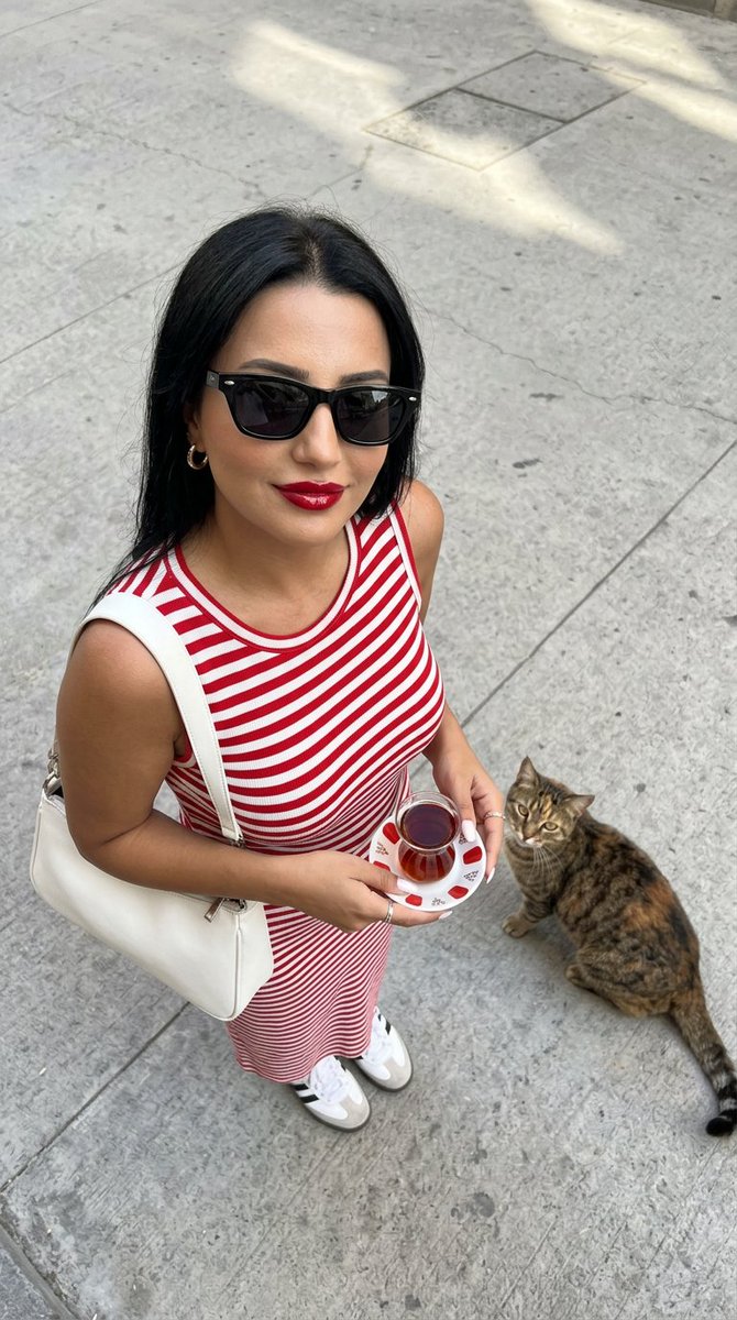 一张高角度俯视的超写实时尚摄影照片：一位时尚年轻女性站立在干净浅灰色的水泥街道上，自信而有魅力地抬头直视镜头。她留着没有头巾的深色长发，唇色为光泽的樱桃红，拥有温暖的古铜色肌肤，并佩戴着细长的...