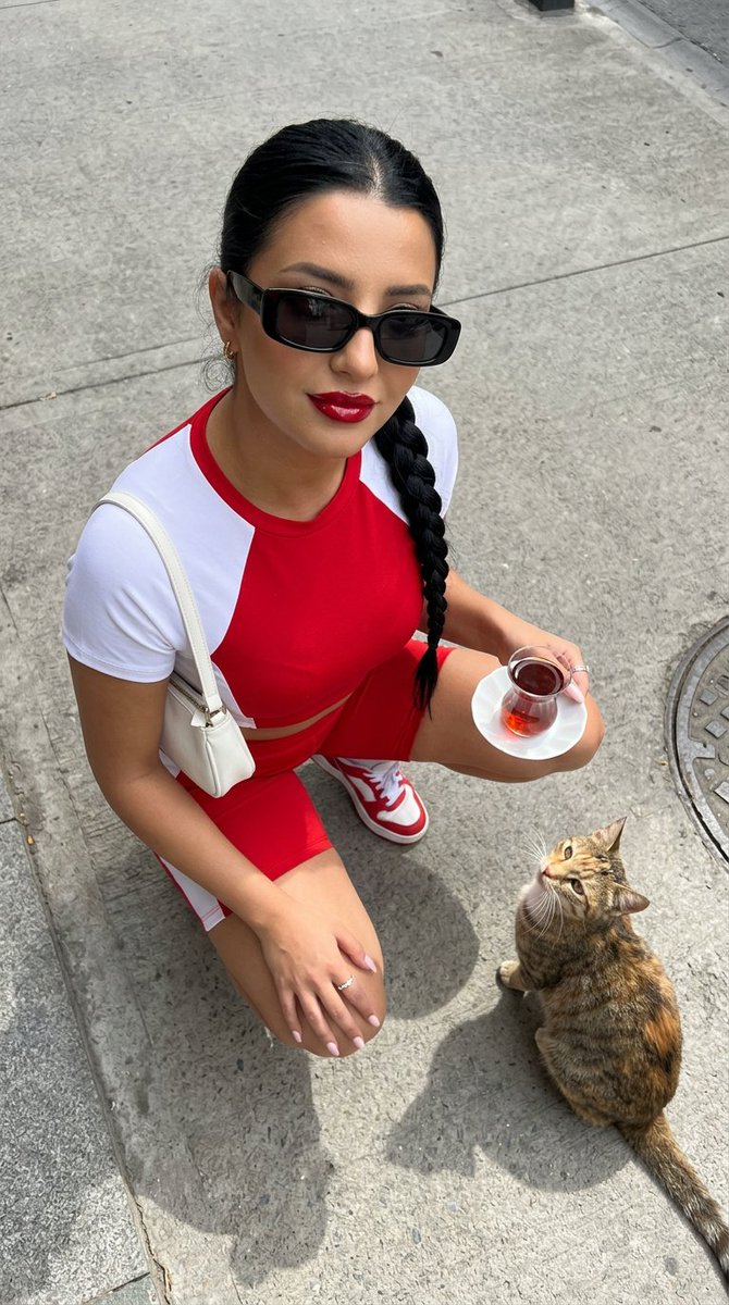 一张高角度俯视的超写实时尚摄影照片：一位时尚年轻女性站立在干净浅灰色的水泥街道上，自信而有魅力地抬头直视镜头。她留着没有头巾的深色长发，唇色为光泽的樱桃红，拥有温暖的古铜色肌肤，并佩戴着细长的...
