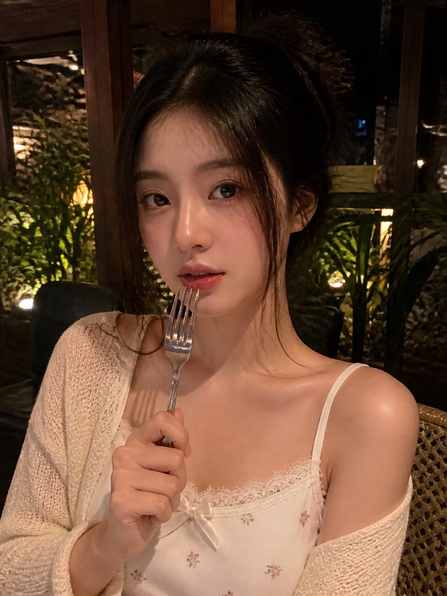 一张写实风格的弱光手机人像照片，主体是一位年轻成年女性，夜晚坐在一家温馨的咖啡馆或餐厅内。近景半身构图，画面比例约为 4:5 或接近正方形。相机举在胸口到脸部之间的高度，从近距离平视或略高于平...