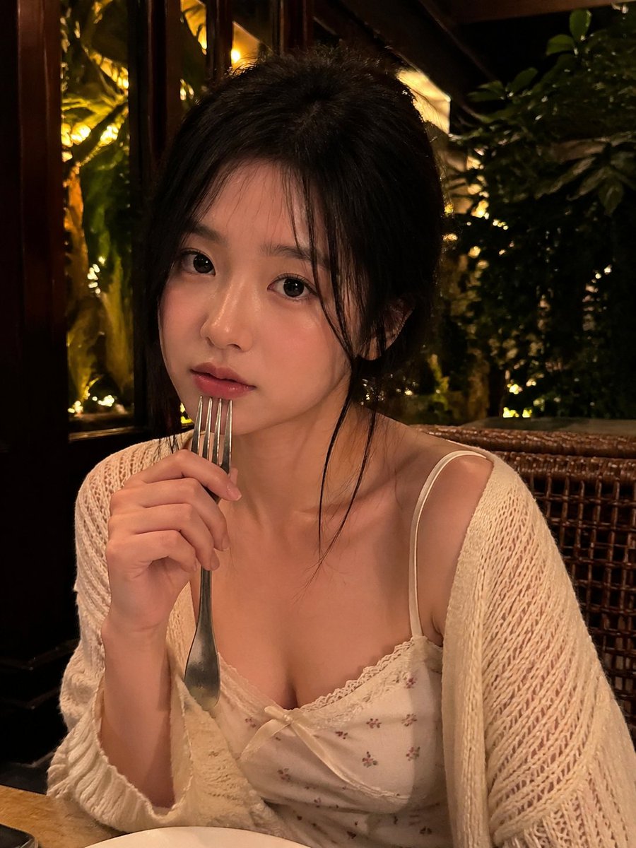 一张写实风格的弱光手机人像照片，主体是一位年轻成年女性，夜晚坐在一家温馨的咖啡馆或餐厅内。近景半身构图，画面比例约为 4:5 或接近正方形。相机举在胸口到脸部之间的高度，从近距离平视或略高于平...