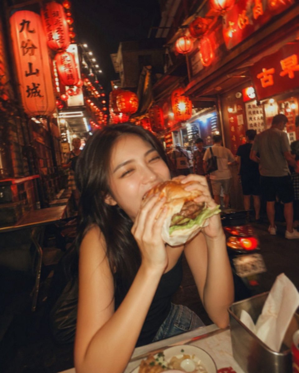 失焦的业余快照：一位亚洲超级模特站在夜晚的老台湾街道中央，发光的红灯笼照亮了整个场景。她坐在街边小吃桌前，吃着一个多汁的大汉堡，心情愉悦。暖色调。Pinterest 美学。JPG。 #AIar...