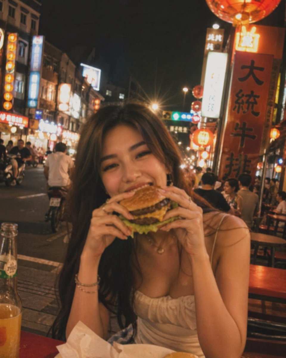 失焦的业余快照：一位亚洲超级模特站在夜晚的老台湾街道中央，发光的红灯笼照亮了整个场景。她坐在街边小吃桌前，吃着一个多汁的大汉堡，心情愉悦。暖色调。Pinterest 美学。JPG。 #AIar...