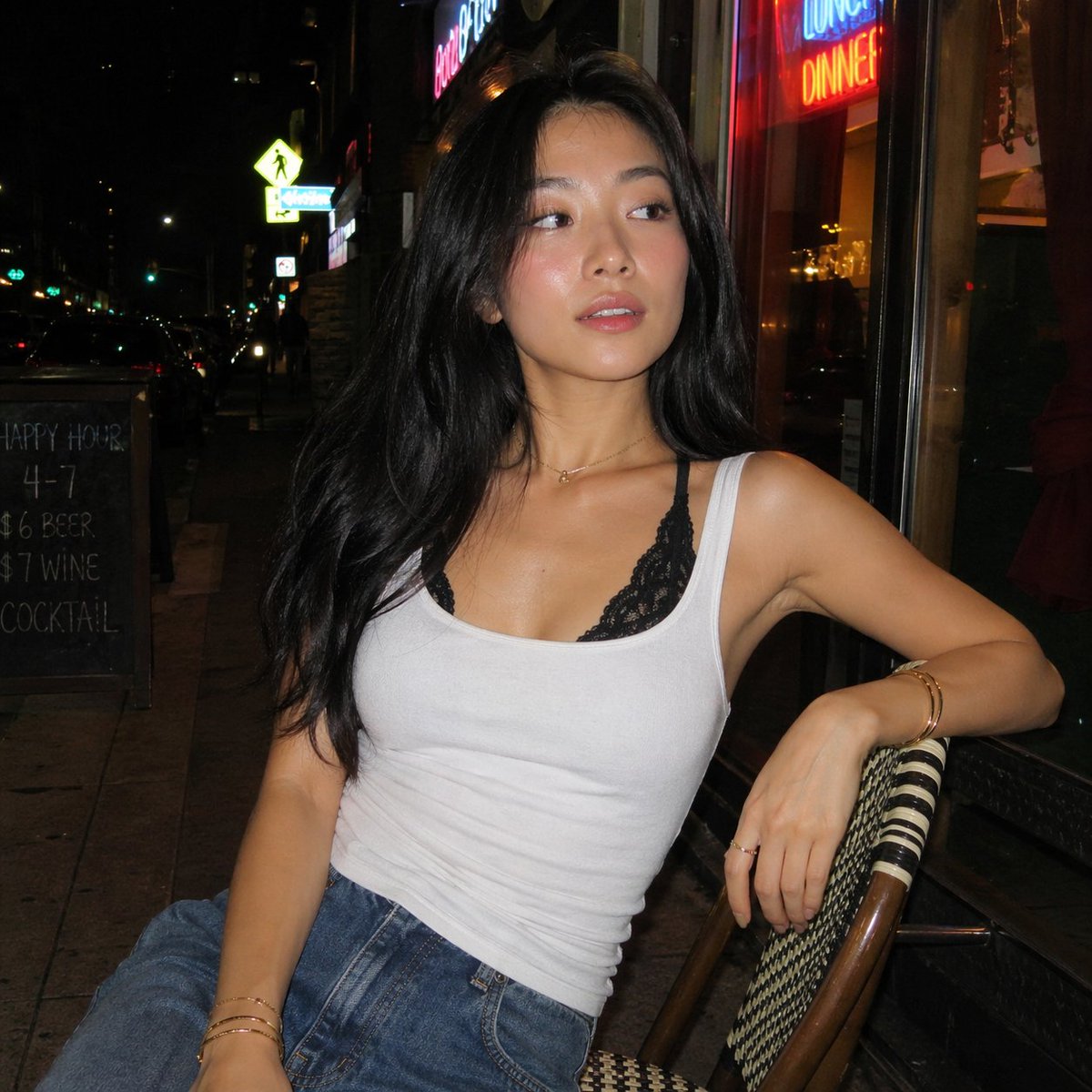 一张夜晚街头的随性人像照，照片中一位年轻亚洲女性随意地坐在一家小型城市餐厅外的藤编咖啡椅上。她有着黑色长发，妆容自然并带有柔和的光泽，身穿白色背心，内搭黑色蕾丝胸衣，下身搭配宽松的牛仔牛仔裤。...