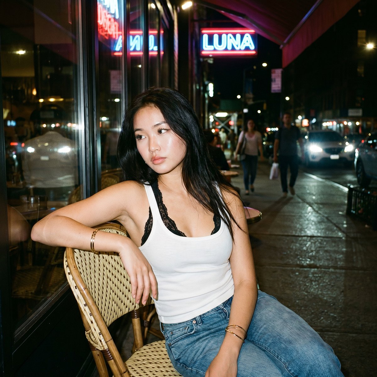 一张夜晚街头的随性人像照，照片中一位年轻亚洲女性随意地坐在一家小型城市餐厅外的藤编咖啡椅上。她有着黑色长发，妆容自然并带有柔和的光泽，身穿白色背心，内搭黑色蕾丝胸衣，下身搭配宽松的牛仔牛仔裤。...