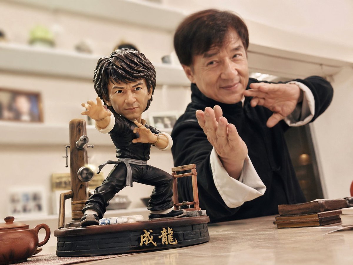 以手机随手拍摄的日常风格，桌面上摆放着一款 【成龙】【功夫】 动漫手办，动作夸张帅气，装备齐全。 同时，真实世界的对应人物也出现在镜头中，与手办...