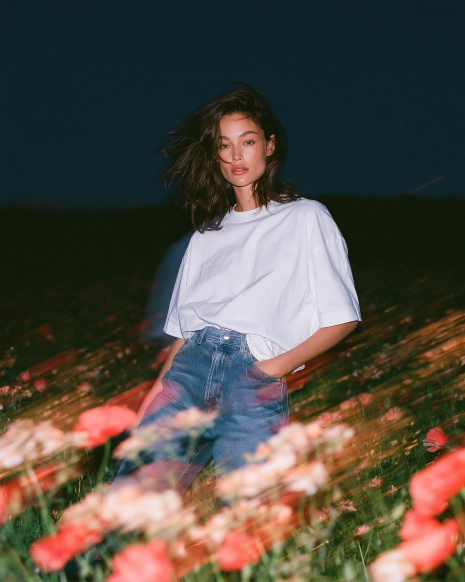 写实风格的实验性时尚人像：一位天生丽质的年轻女子正面站立于蓝色时刻狂风拂过的野花田中，镜头从大腿向上以平视取景。她身穿一件宽松的纯白棉质T恤，搭配水洗中蓝色直筒牛仔裤，日常随性的自然穿搭，深色...