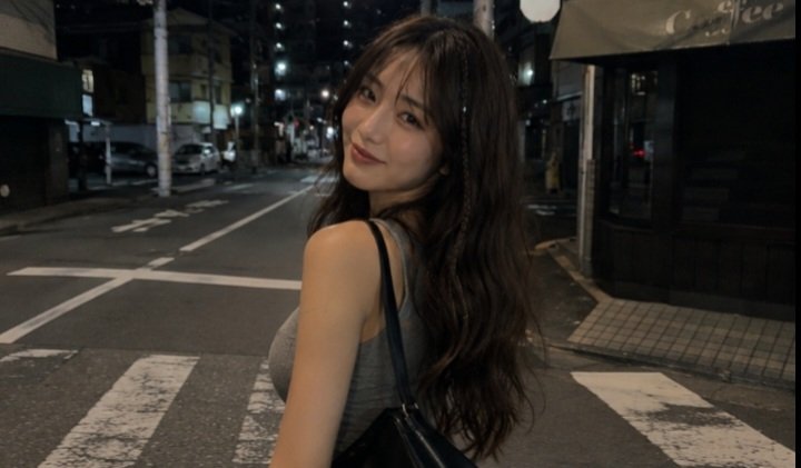 一张电影感十足的夜间街头抓拍人像，拍摄于日本街头，使用 iPhone 拍摄。一名年轻女子，长长的深棕色波浪卷发中编有一缕细辫，淡妆，嘴角带着若有若无的微笑，回头望向镜头。她身穿灰色无袖上衣，搭...