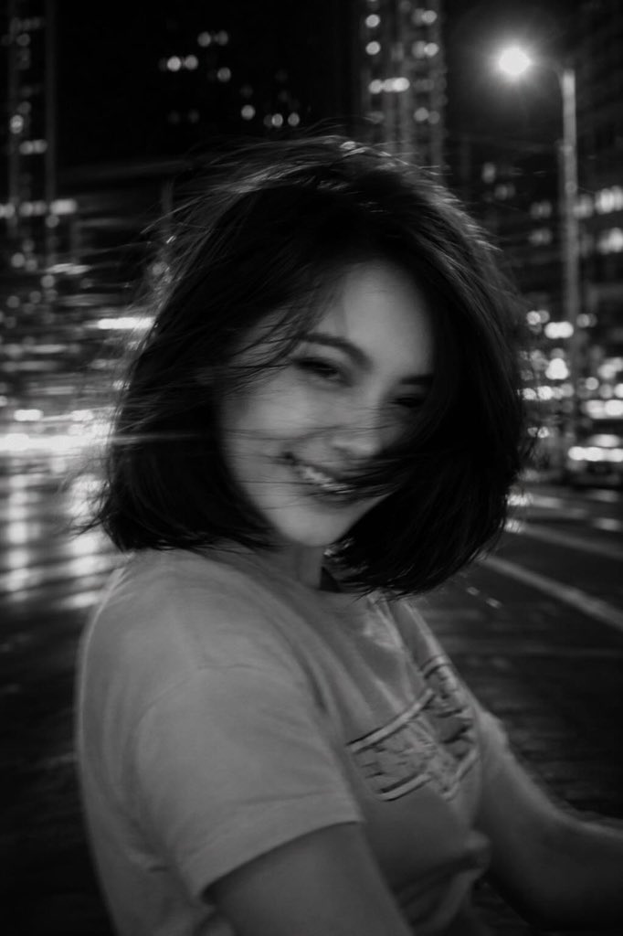 一张黑白电影感的城市夜景照片，画面中一位年轻女子位于前景，略微偏离中心，正在转头过程中，长发在画面中甩动成柔和、幽灵般的发丝。长曝光拍摄，带有意运动模糊。她面部特征部分模糊但仍可辨认——富有表...