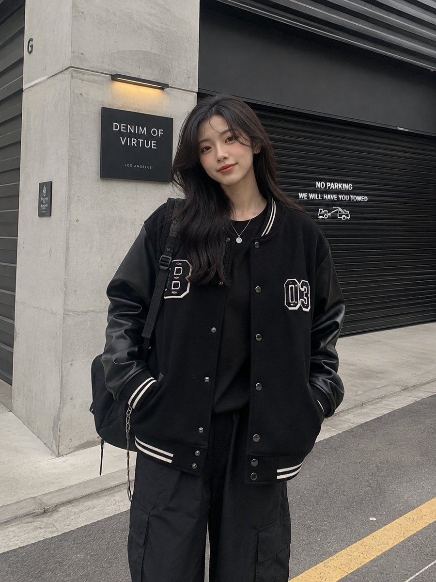 一张潮流街头服饰时尚人像照：一位时尚年轻女性自信地站在一座现代工业建筑前，建筑拥有混凝土墙面和一扇黑色金属卷帘门。她身穿一件超大版型的黑色棒球外...
