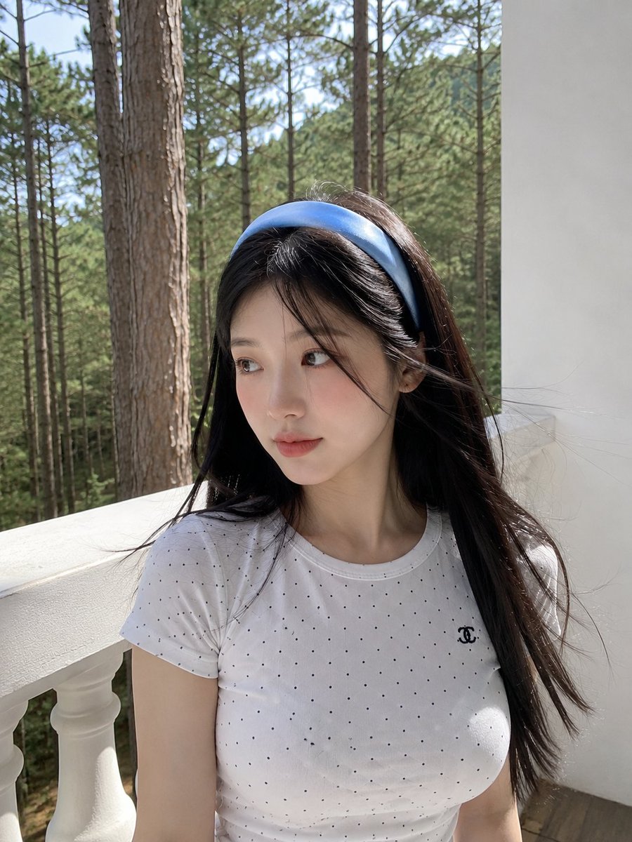 超写实的智能手机照片：一位美丽的年轻日本女性站在被宁静松林环绕的白色阳台上。她拥有白皙的瓷器肌肤、柔和的玫瑰色脸颊、深棕色的大眼睛、精致细腻的五官，以及在微风中轻轻飘动的黑色长直发。她佩戴着一...