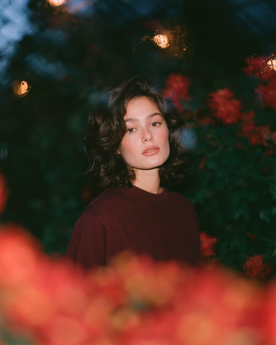 梦幻的电影感夜景人像：一位美丽的年轻女性，留着短款波浪深色头发，皮肤散发着自然光泽，身穿一件宽松的酒红色圆领卫衣，夜晚站在一座郁郁葱葱的花园中，园内开满了鲜艳的红色花朵和深绿色的枝叶。镜头透过...
