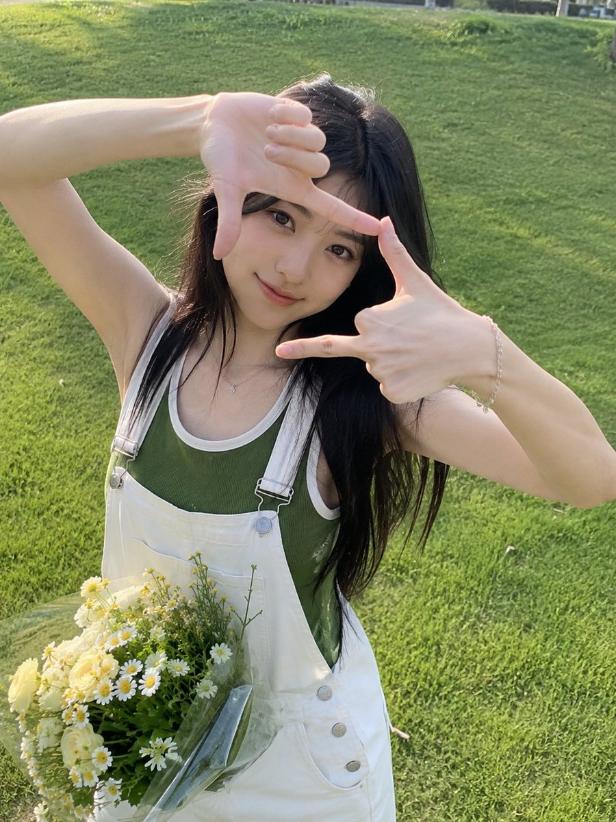 一张迷人的户外人像照,一位年轻女子站在郁郁葱葱的绿色草坪上,直视镜头,双手俏皮地在脸部周围比划出一个长方形相框。她有着一头长而深色、柔和波浪卷的...