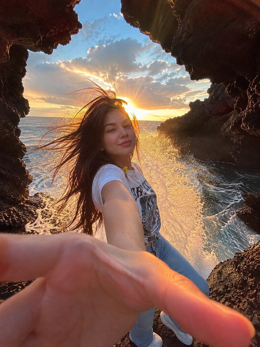 一幅电影感十足的海边人像照：一位年轻女子站在天然海洞内的黑色火山岩上，洞口粗犷的岩石形成天然画框。她一头深色长发在海风中戏剧性地飘扬，对着镜头俏皮地眨着一只眼，嘴角带着温柔微笑。她身穿一件休闲...