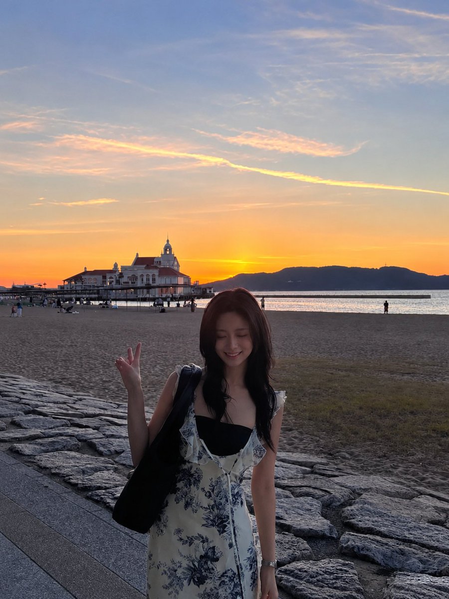 超现实电影感旅行照片：一位年轻的东亚女性站在沙滩上，沐浴在令人屏息的日落中，采用竖向 3:4 构图。她身穿一件精致的白色连衣裙，点缀着细腻的蓝色花卉印花，肩带为荷叶边设计，整体剪裁贴合身形。她...