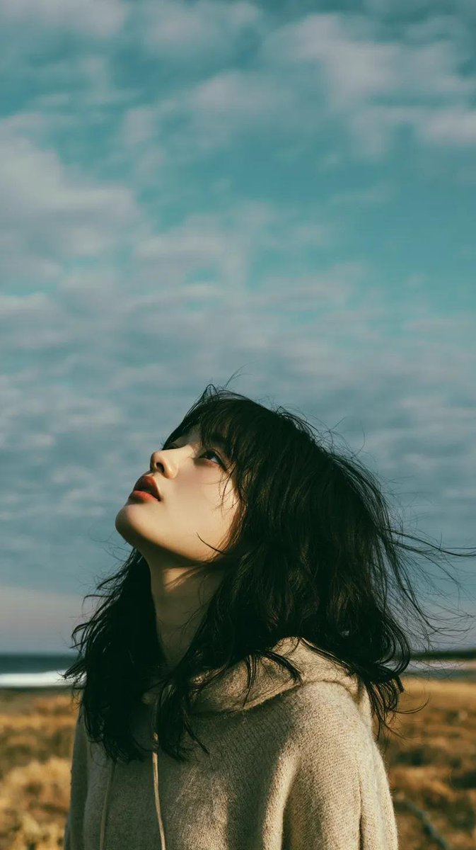 电影极简风格人像：一位美丽的年轻东亚女性独自站立在一片开阔的野花草地上，头顶是无尽的蓝天，她微微仰头，双眼或闭或半睁，仿佛在拥抱风。黑色长发自然蓬松、随风自由拂过面庞，肌肤柔润如瓷，五官精致，...
