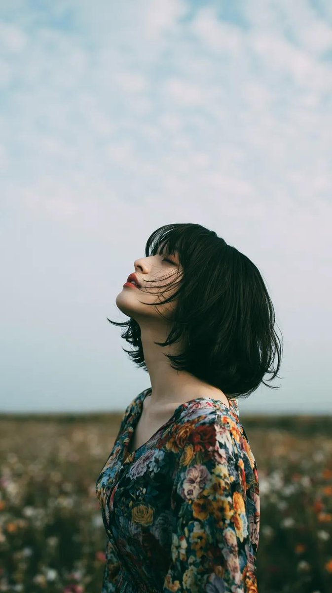 电影极简风格人像：一位美丽的年轻东亚女性独自站立在一片开阔的野花草地上，头顶是无尽的蓝天，她微微仰头，双眼或闭或半睁，仿佛在拥抱风。黑色长发自然蓬松、随风自由拂过面庞，肌肤柔润如瓷，五官精致，...