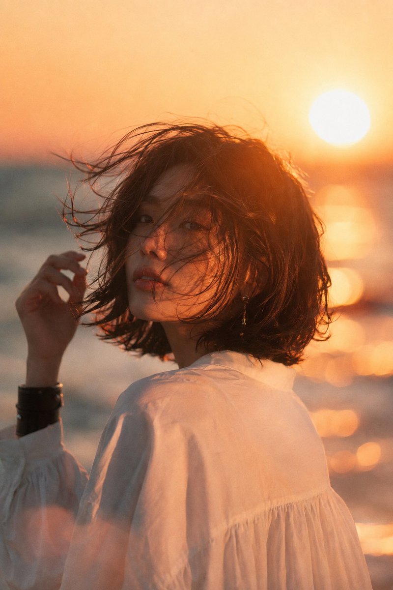海滩日落中的美丽年轻亚洲女性，中景电影肖像。深色短发波波头在海风中飘扬，头部微向左倾，手正在整理头发。甜美浪漫的瞬间。精致优雅的耳饰，简约宽松的白色衬衫，左腕佩戴黑色宽手镯。柔和自然的妆容，沉...