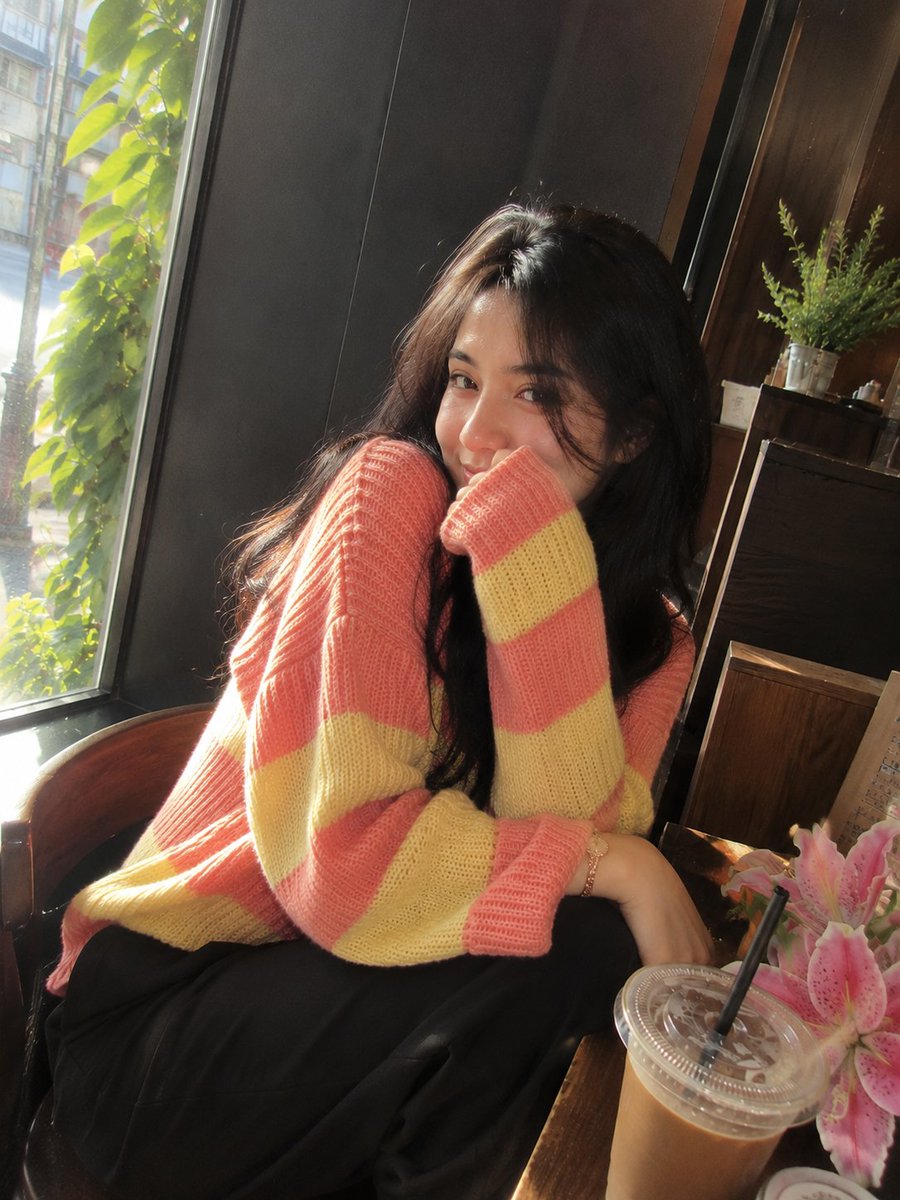 照片级写实风格的影院感咖啡馆肖像：一位美丽的成年女性，留着长而自然卷曲的深棕黑色头发，皮肤柔和泛光，五官精致，眼神富有表现力。她身穿宽松舒适的针织毛衣，交替呈现柔和的粉色与浅黄色横向条纹，搭配...